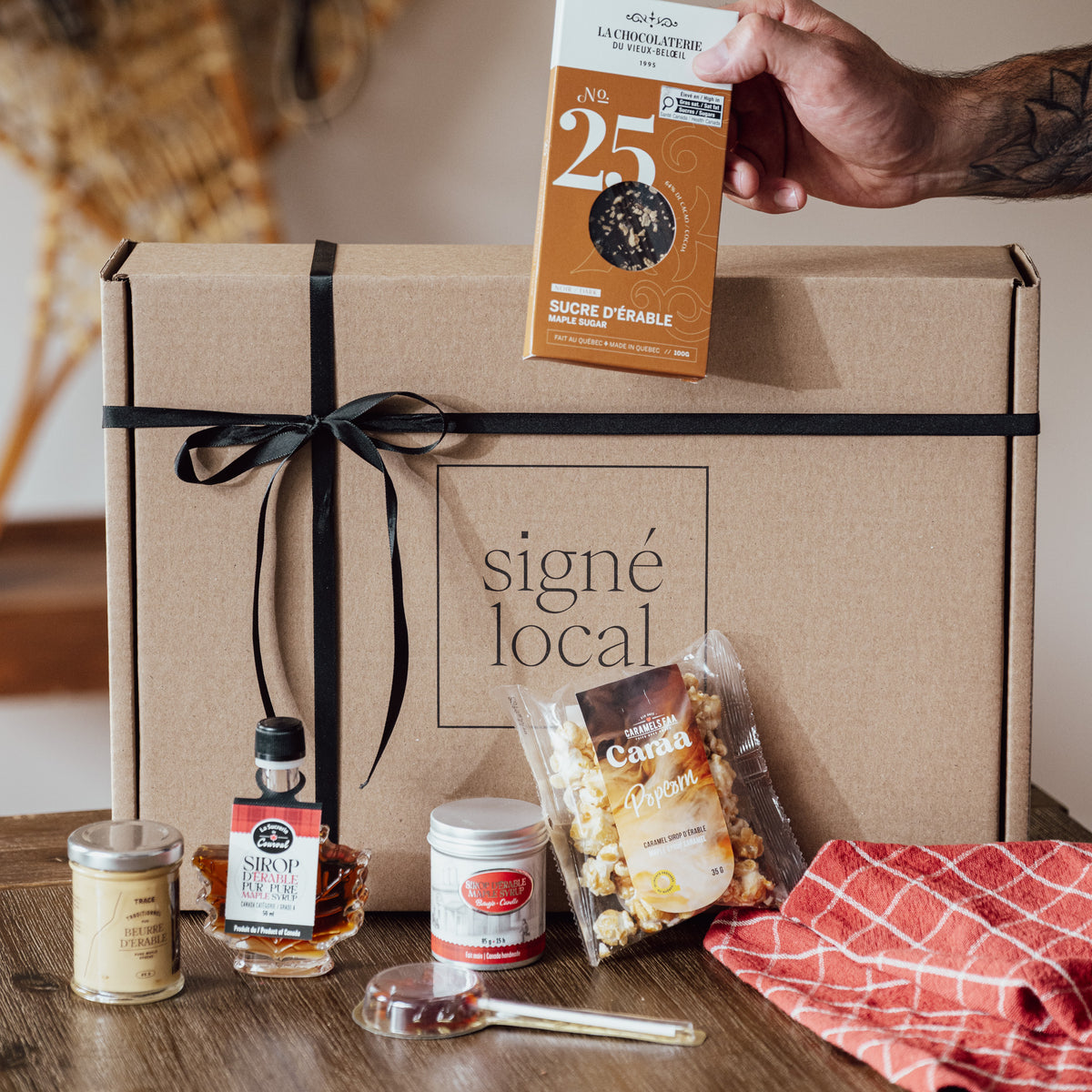Coffret Comme à la cabane Signé Local en situation : chocolat au sucre d'érable de La Chocolaterie du Vieux-Beloeil, sirop d'érable, beurre d'érable TRACE, popcorn au caramel et suçon d'érable devant une boîte kraft au ruban noir