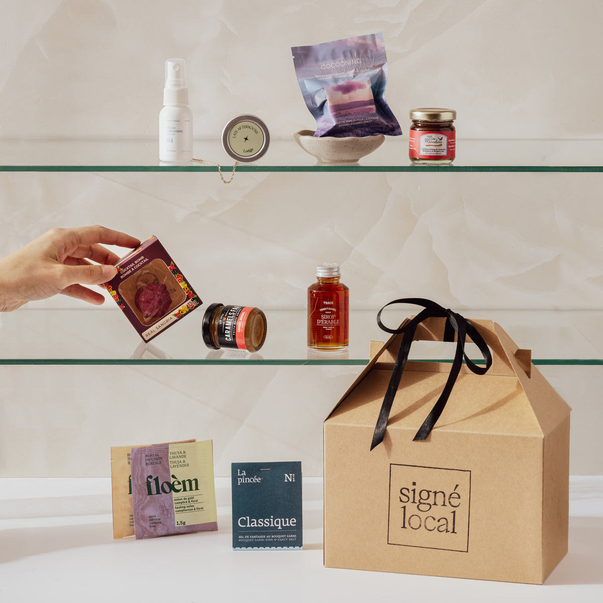 Coffret Les Meilleurs en Minis Signé Local en situation : boîte à pignon kraft et formats minis sur des tablettes de salle de bain, infusions Floèm, épices La Pincée, sirop d'érable, bombe à cocktail et brume